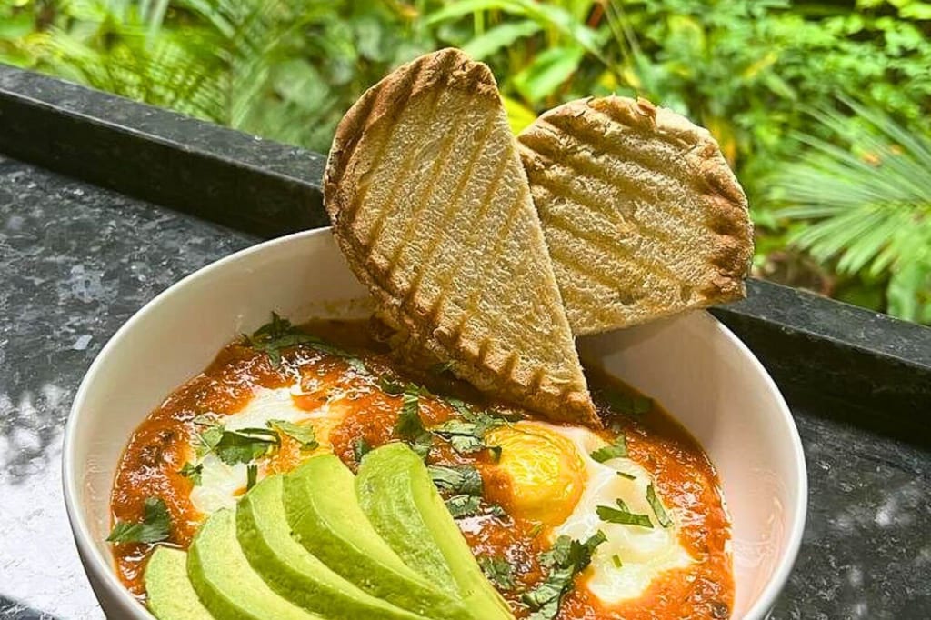 Schüssel mit Eiern in Tomatensauce, Avocadoscheiben und zwei gerösteten Fladenbroten auf einem Balkon.