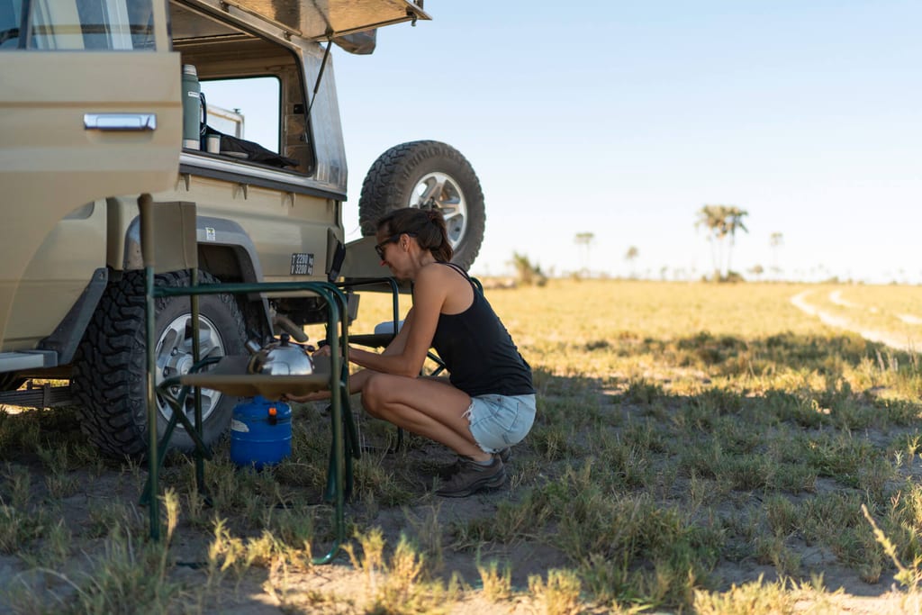 Frau richtet Campingkocher neben Geländewagen auf offener Graslandschaft bei hellem Tageslicht