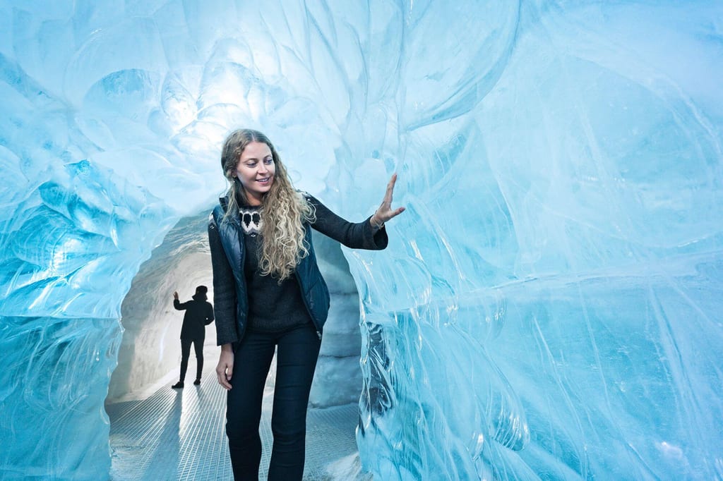 Zwei Personen in einer blau leuchtenden Eishöhle, Frau berührt glatte Eiswand, Silhouette im Hintergrund.