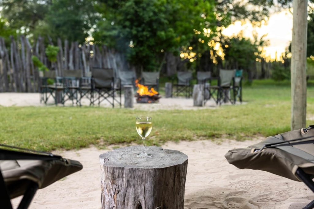 Weinglas auf Holzstamm vor Feuerstelle und Klappstühlen in einem Campinglager bei Abendlicht