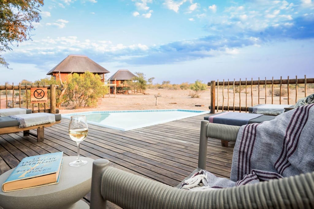 Holzterrasse mit Pool, Weinglas und Blick auf strohgedeckte Hütten in trockener Savannenlandschaft.