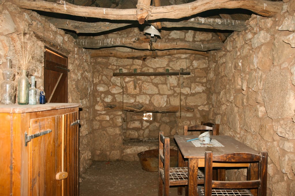 Rustikaler Innenraum einer Steinhütte mit Holztisch, Stühlen und Kommode, warme Beleuchtung.