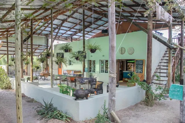 Überdachte Terrasse mit Tischen, hängenden Pflanzen und Außentreppe vor einem pastellgrünen Gebäude.