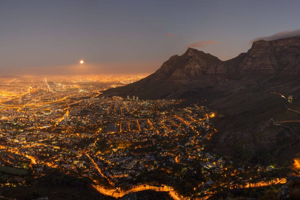Panoramablick auf eine nachts beleuchtete Stadt mit dunkler Bergsilhouette und Mond am Abendhimmel.