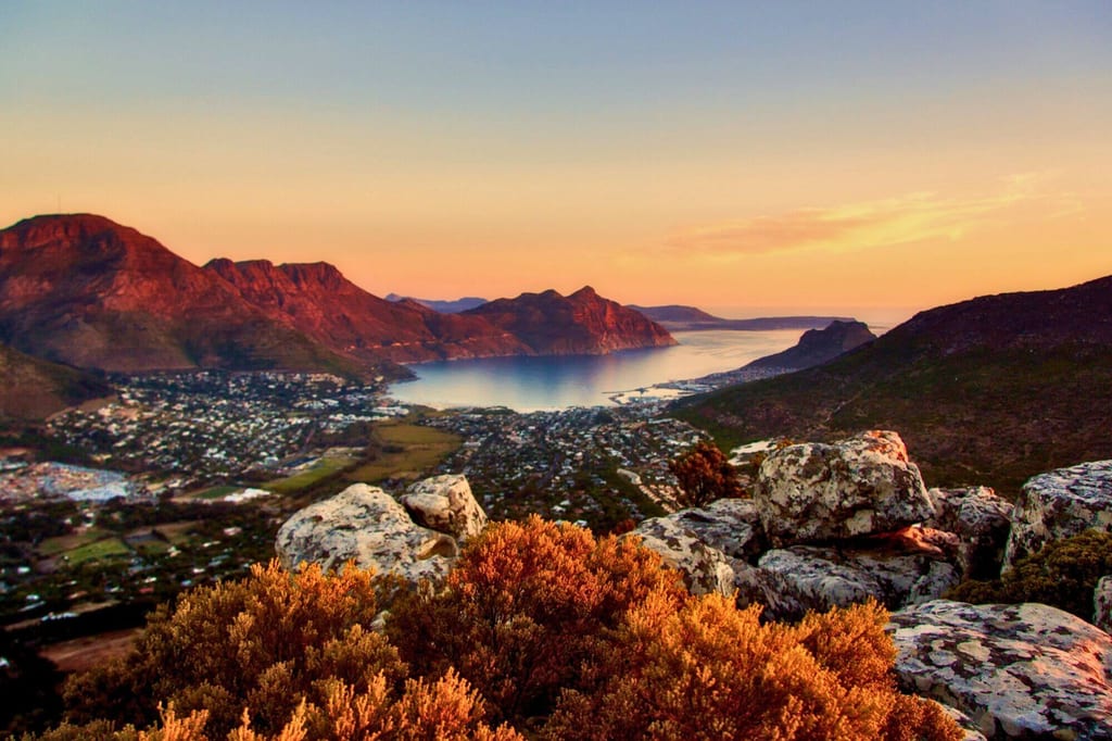 Felsiger Hang mit Buschwerk im Vordergrund, Blick auf eine Küstenbucht und Berge bei Sonnenuntergang im Western Cape, Südafrika.