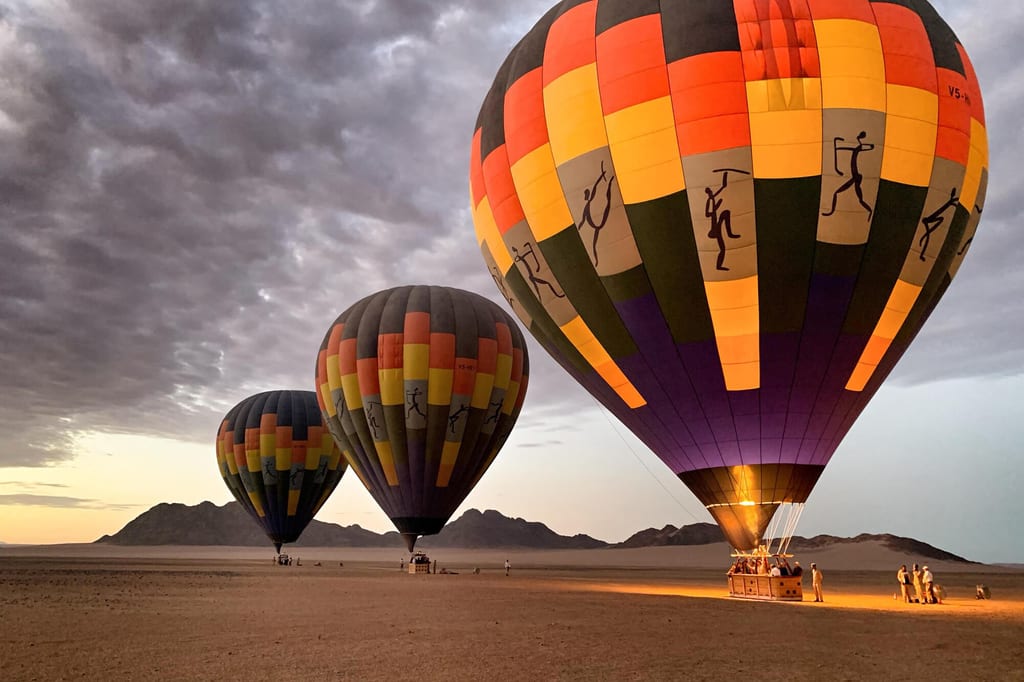 Drei bunte Heißluftballons auf einer Wüstenebene bei Dämmerung, Personen an den Körben, warme Brennerlichter