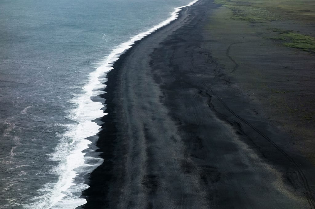 Luftaufnahme einer langen schwarzen Sandküste mit ziehenden Wellen und spärlicher Vegetation