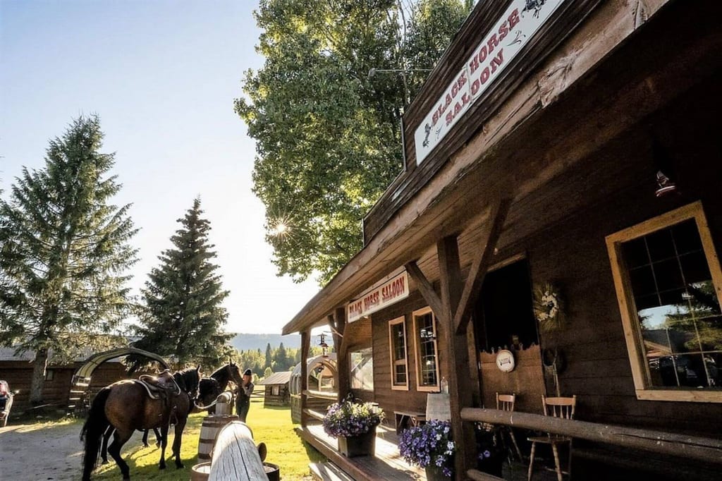 Westlicher Saloon-Holzbau mit Veranda, zwei gesattelte Pferde und sonnige ländliche Szenerie