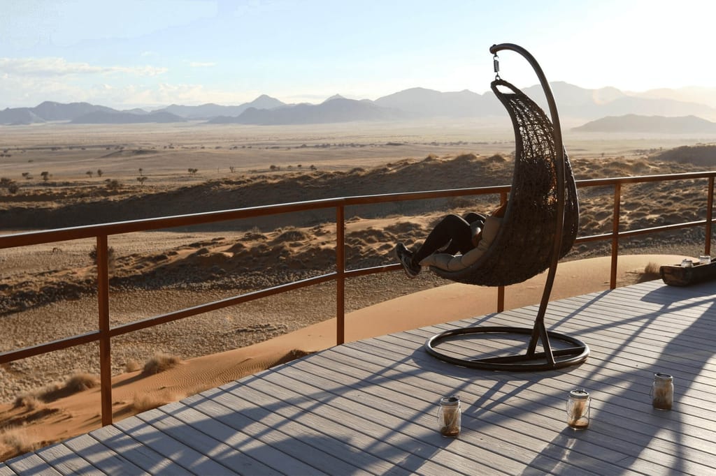 Person in Hängesessel auf Terrasse blickt bei warmem Nachmittagslicht über weite Sanddünen und entfernte Berge.