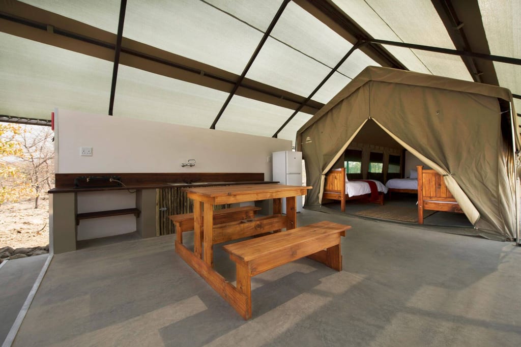 Offene Safari-Zelt-Lodge mit Holztisch, Bänken und zwei Einzelbetten im Inneren.