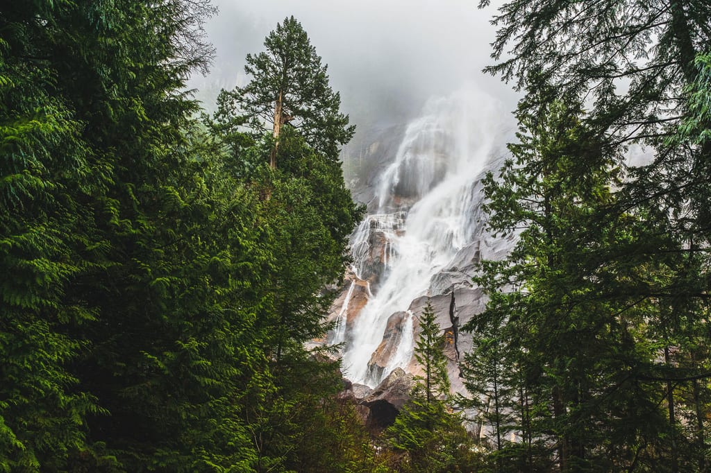 Wasserfall, eingerahmt von dichtem Nadelwald bei bewölktem Tageslicht