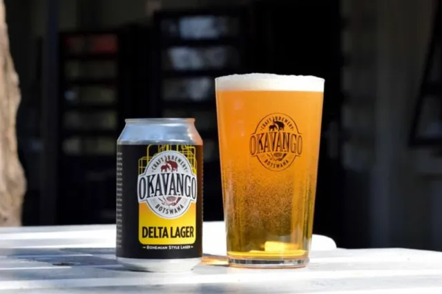 Bierdose und Bierglas mit Okavango Delta Lager auf hellem Holztisch bei Tageslicht.