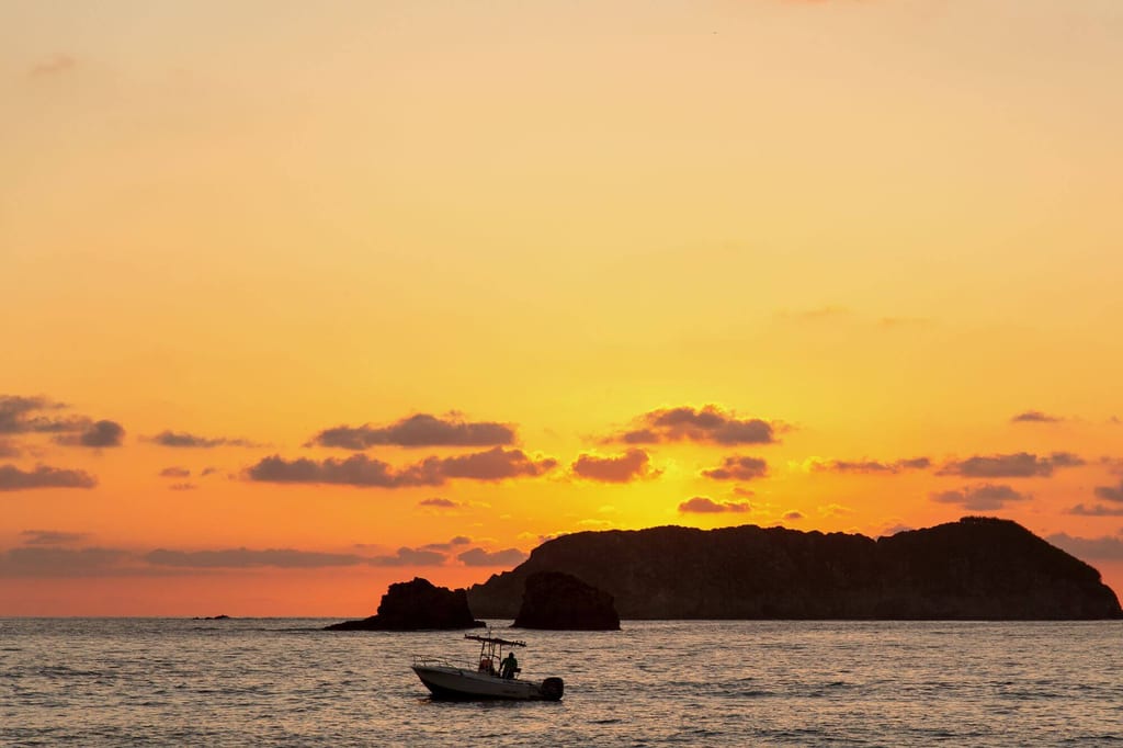 Motorboot vor felsiger Insel bei orangefarbenem Sonnenuntergang über ruhigem Meer.