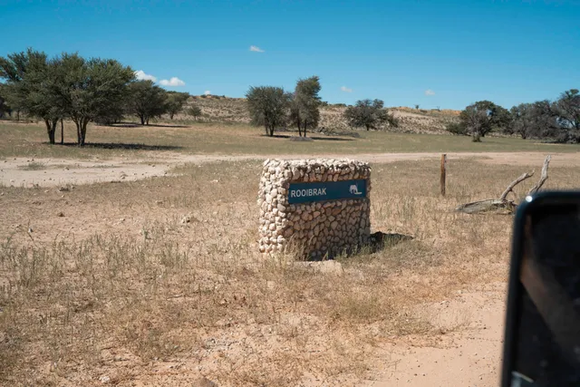 Steinschild mit Aufschrift ‚ROOIBRAK‘ in trockener, baumbestandener Savannenlandschaft bei klarem Himmel.