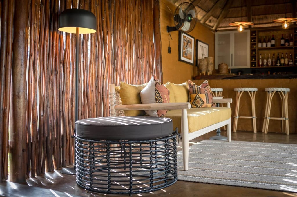 Gemütliche Lounge mit gelbem Sofa, Geflecht-Hocker und Holzwand bei warmem Tageslicht.