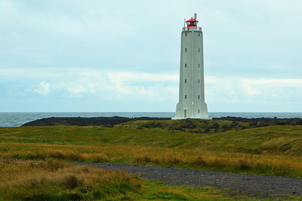 Weißer Leuchtturm an einer grasigen Küste bei bewölktem Tageslicht, frontal aus der Entfernung.