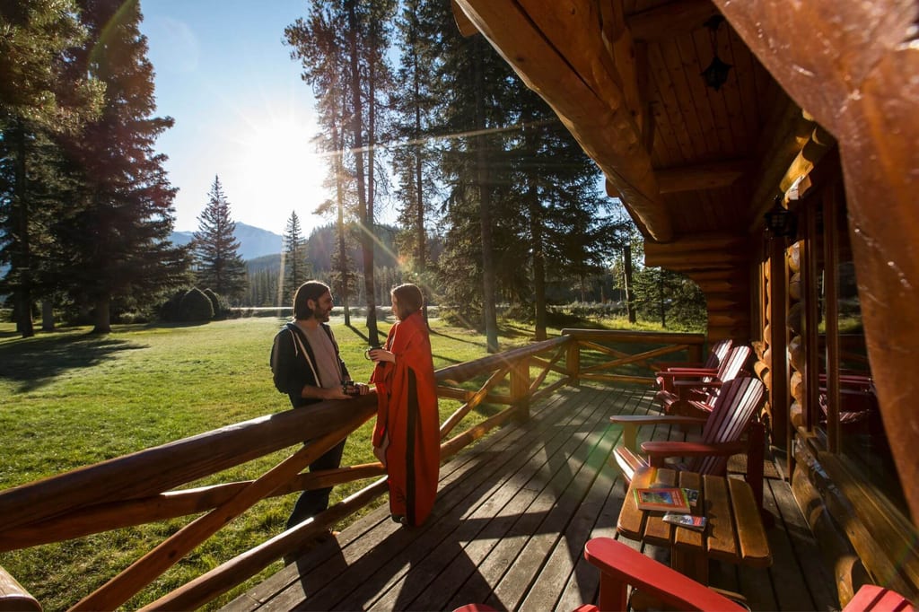Zwei Personen auf Holzterrasse einer Blockhütte bei untergehender Sonne, Nadelbäume und Berg im Hintergrund.
