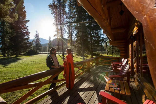 Zwei Personen auf Holzterrasse einer Blockhütte bei untergehender Sonne, Nadelbäume und Berg im Hintergrund.