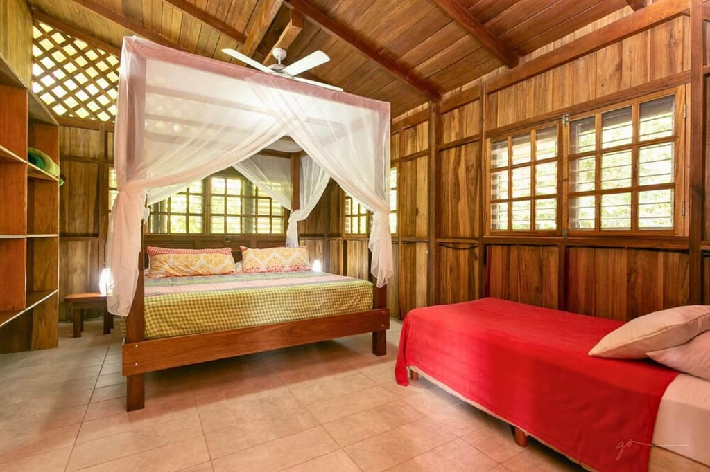 Rustikales Holzschlafzimmer mit Himmelbett und Moskitonetz, Einzelbett mit roter Decke und Sprossenfenstern.