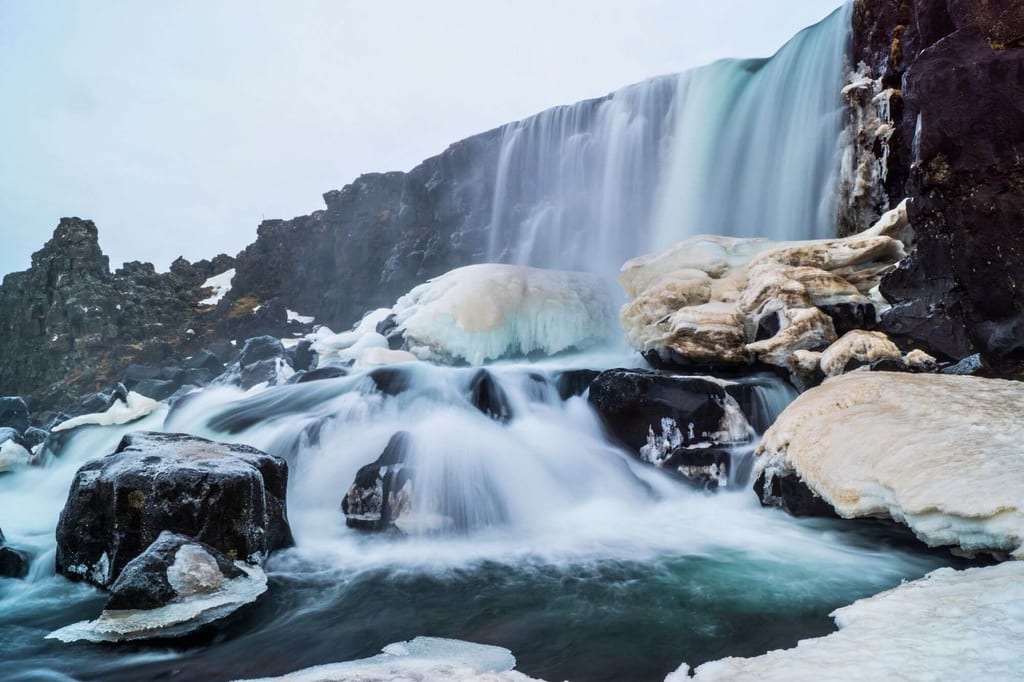 Winterlicher Wasserfall mit eisigen Uferpartien und seidig fließendem Wasser, bewölkter Himmel.
