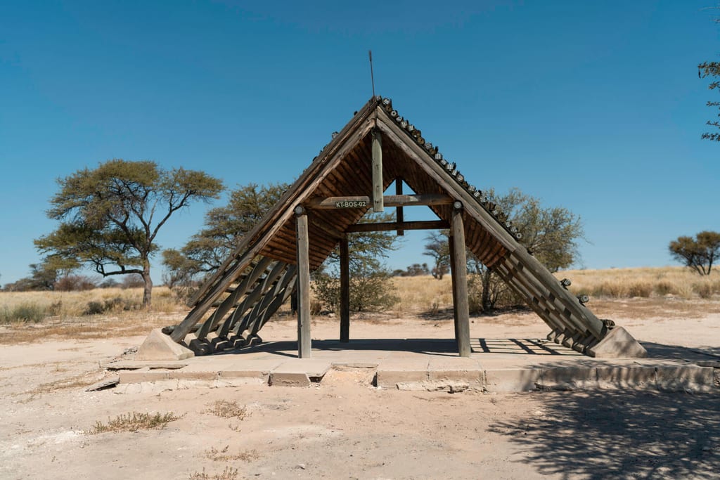 Dreieckige Holzhütte auf Betonplattform in trockener Savannenlandschaft vor blauem Himmel