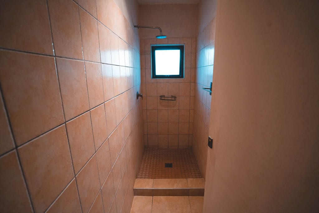 Schmale Dusche mit beigen Fliesen, Duschkopf und kleinem Fenster, Blick von der Tür