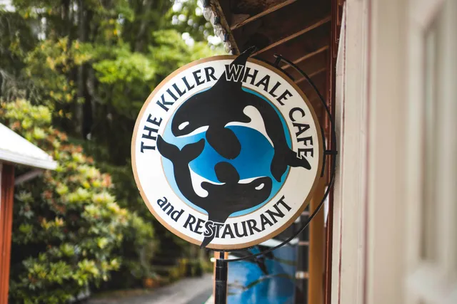 Rundes Caféschild mit stilisierten Killerwalen an einer Gebäudeecke, unscharfer grüner Hintergrund