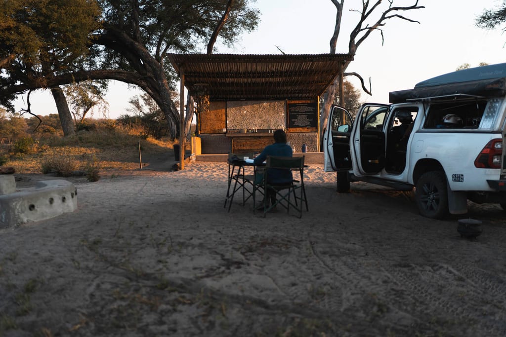 Person sitzt am Camp-Tisch vor offener Hütte und Geländewagen im warmen Abendlicht