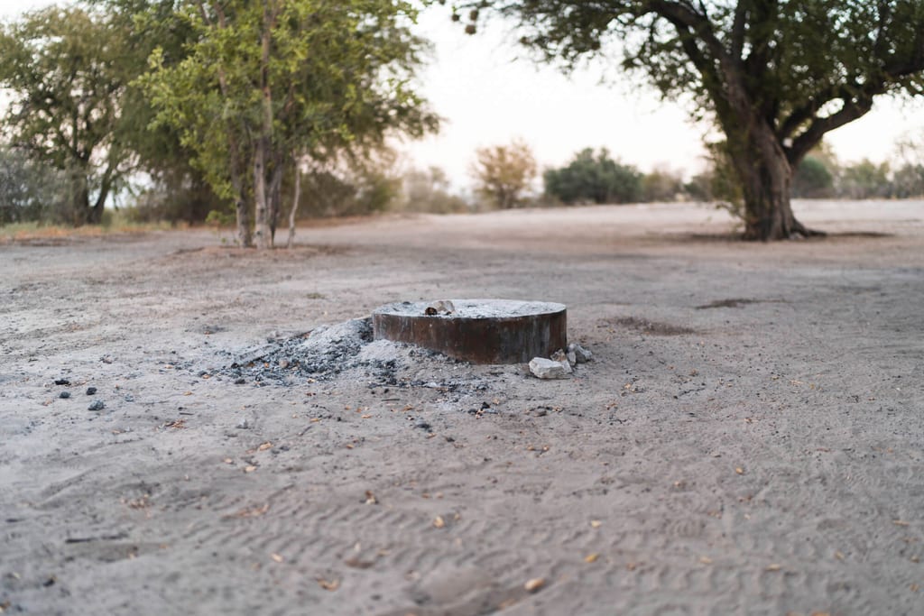 Runde Metallfeuerstelle mit Asche auf sandigem Boden vor vereinzelten Bäumen im weichen Abendlicht.