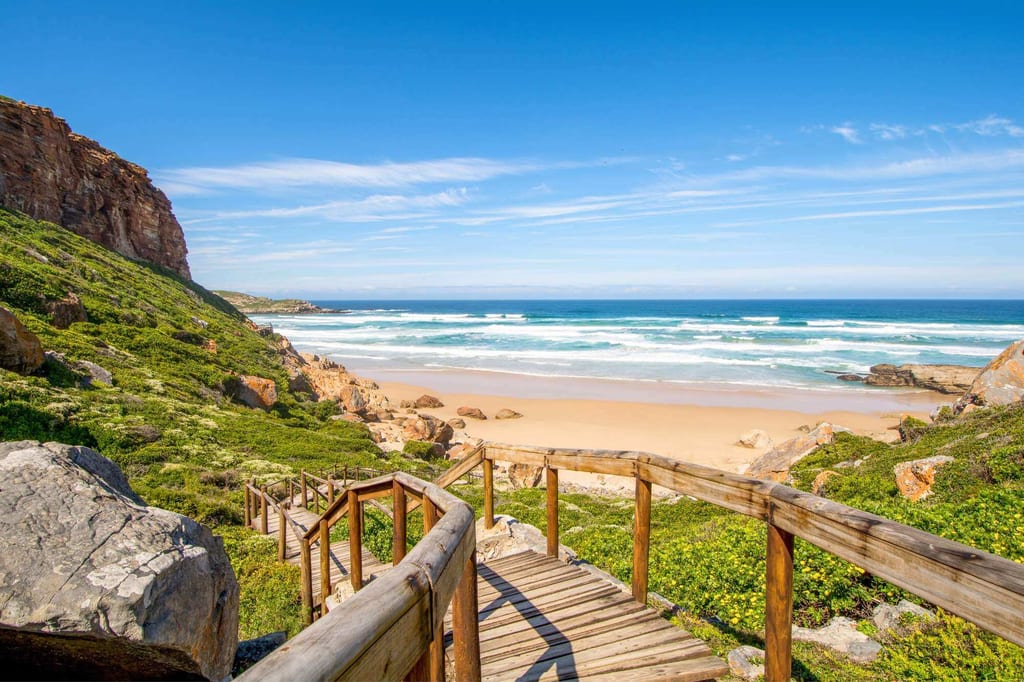 Holzsteg führt die Treppe hinunter zu Sandstrand und blauem Meer an der Garden Route, sonnig und klar.