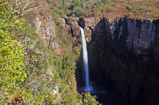 Schmaler Wasserfall stürzt in dunklen Kessel zwischen steilen Felsklippen, Vordergrund mit dichtem Buschwerk