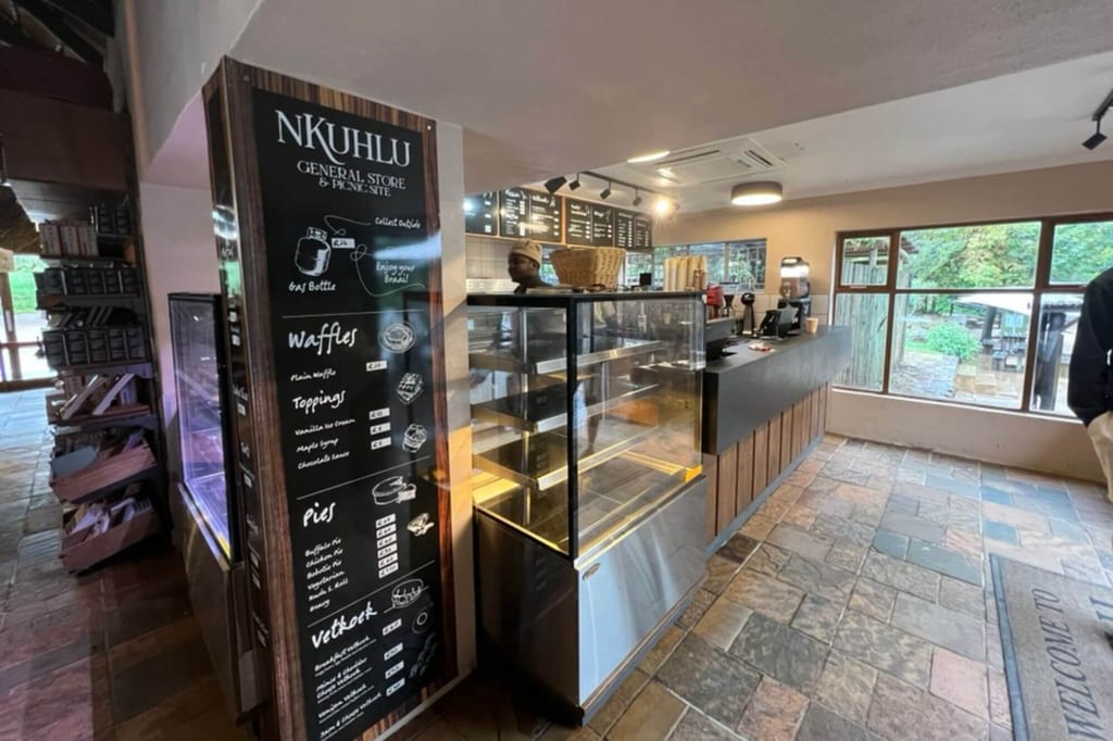 Innenansicht eines kleinen Café-General-Stores mit Theke, Menübrett ‚NKUHLU‘ und großen Fenstern.