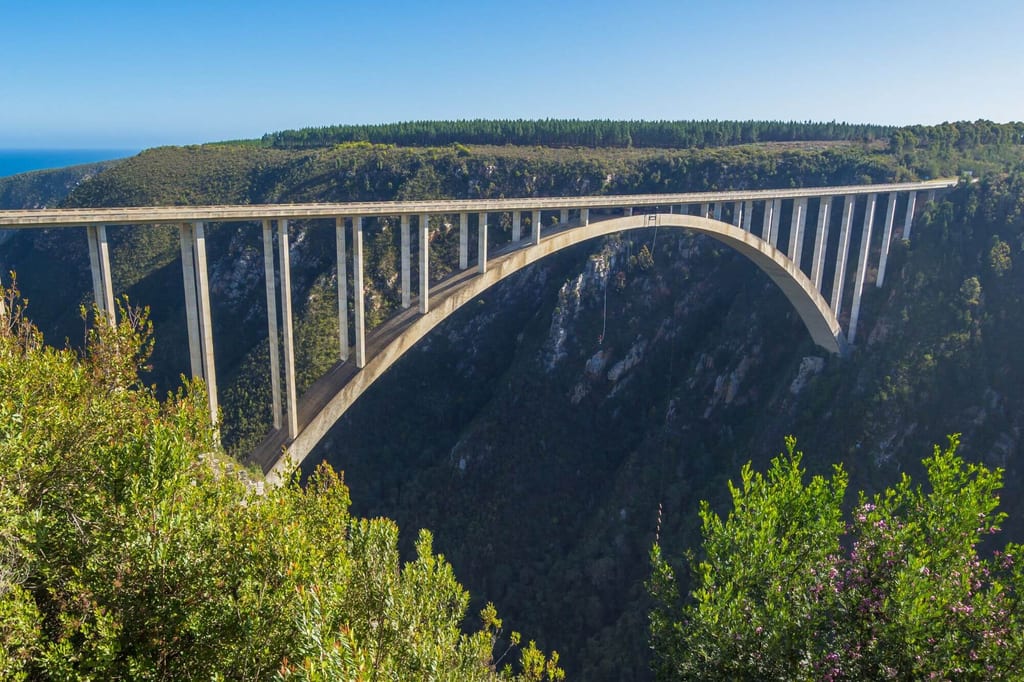 Seitliche Aufnahme einer großen Bogenbrücke über einer bewaldeten Schlucht bei klarem Tageslicht.