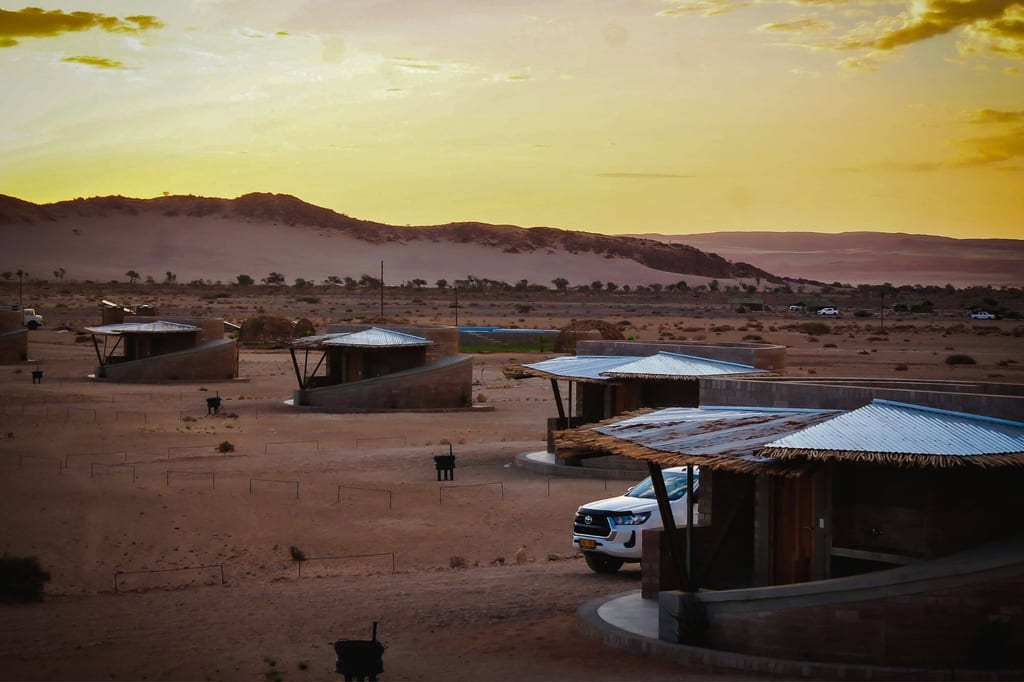 Wüstenanlage mit runden Unterkünften und Dünen im warmen Abendlicht, ruhige Weite bis zum Horizont.