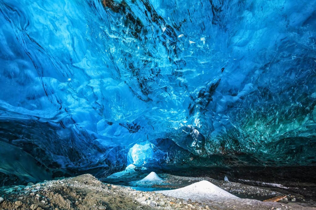 Blick aus der Gletscherhöhle auf leuchtend blaues Eis und kiesigen Boden mit kleinen Eishügeln.