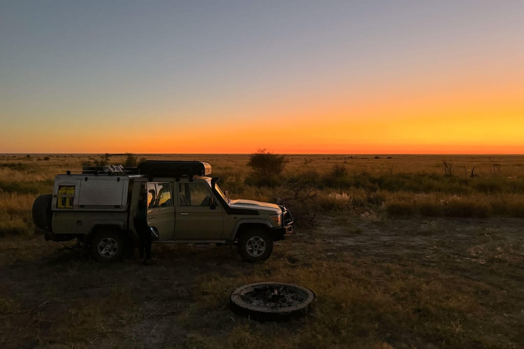 Geländewagen neben Feuerstelle in weiter Savanne bei orangefarbenem Sonnenuntergang.