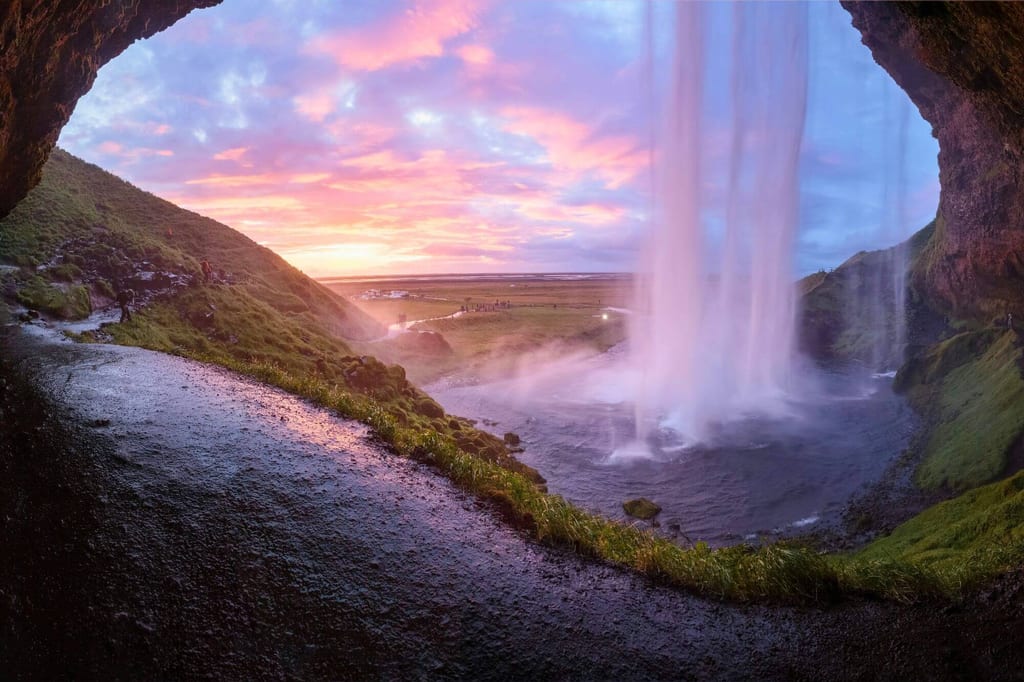 Blick aus einer Höhlenöffnung auf einen Wasserfall und einen rosa-violetten Sonnenuntergang über einer Ebene.