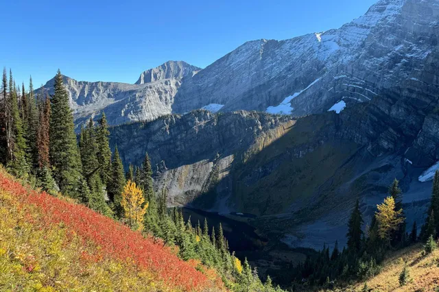 Alpines Bergtal mit bunten Herbsthängen, Nadelwald und kleinem Bergsee unter klar blauem Himmel.