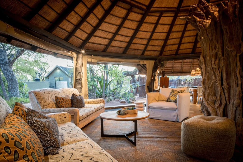 Gemütliche Lodge-Lounge unter Strohdach mit Sofas und Blick in den Garten, warmes Abendlicht.
