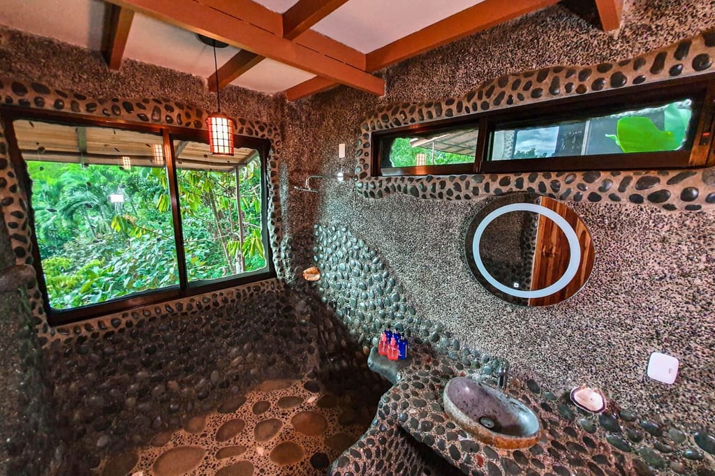 Rustikales Badezimmer mit Flusssteinwänden, großem Fenster und Blick auf dichten Regenwald.