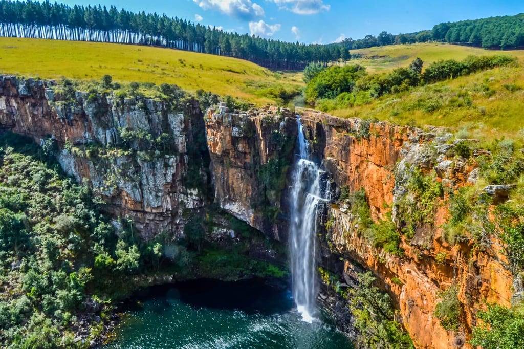 Luftaufnahme eines Wasserfalls, der von einer roten Felsklippe in ein dunkles Becken stürzt.