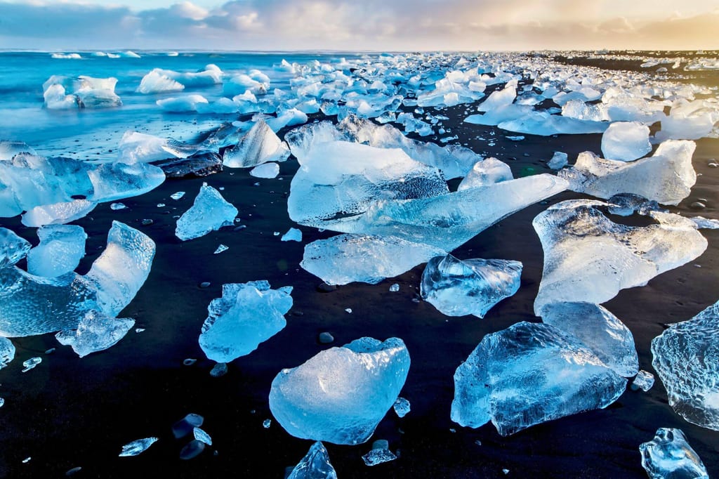 Klare Eisblöcke liegen verstreut auf schwarzem Sandstrand, Meer und warmes Licht am Horizont.