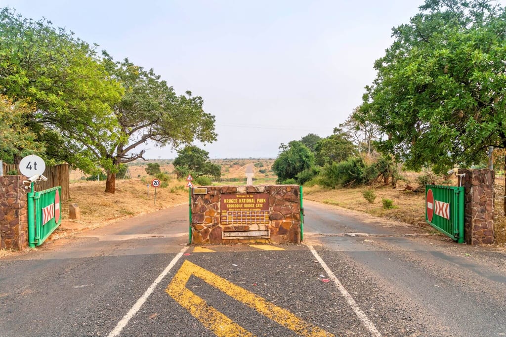 Frontansicht eines Parkeingangs mit Steinbarriere und grünen Toren vor trockener Savannenlandschaft