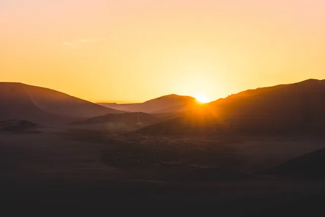 Weite Wüstenlandschaft mit tiefstehender Sonne hinter sanften Dünen, warmes goldenes Morgenlicht.