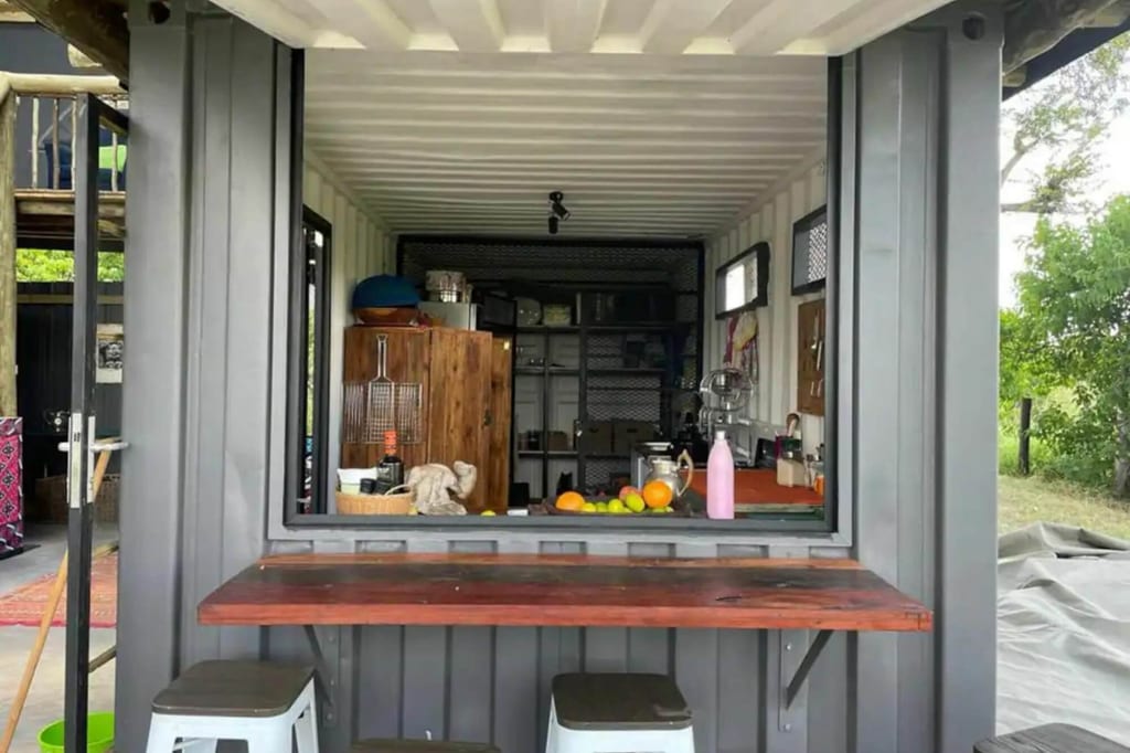 Frontalaufnahme einer offenen Containerküche mit Bartresen, Hockern und Obst auf der Arbeitsfläche.