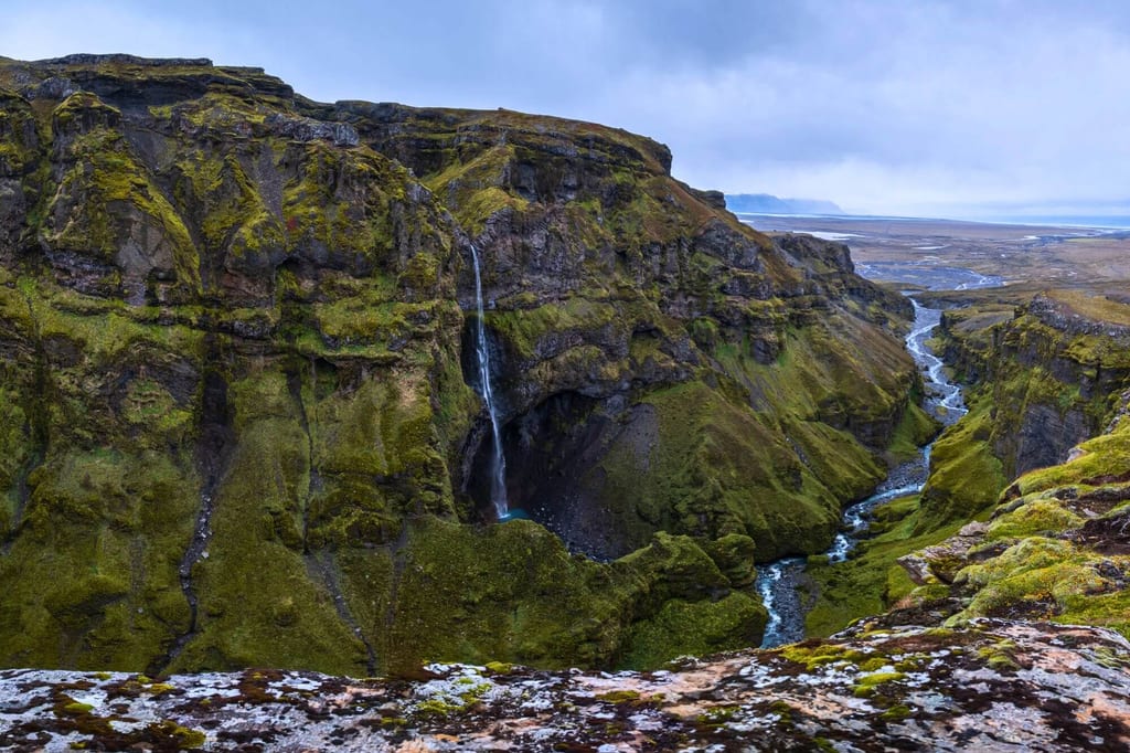 Blick von einem Felsrand in eine moosbedeckte Schlucht mit schmalem Fluss und hohem Wasserfall bei bewölktem Himmel.