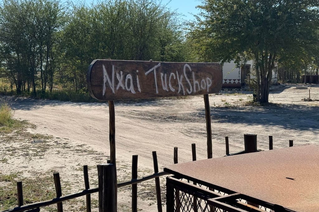 Hölzernes Schild "Nxai TuckShop" über sandiger Straße, Bäume im Hintergrund, sonniger Tag