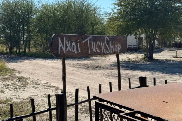 Hölzernes Schild "Nxai TuckShop" über sandiger Straße, Bäume im Hintergrund, sonniger Tag