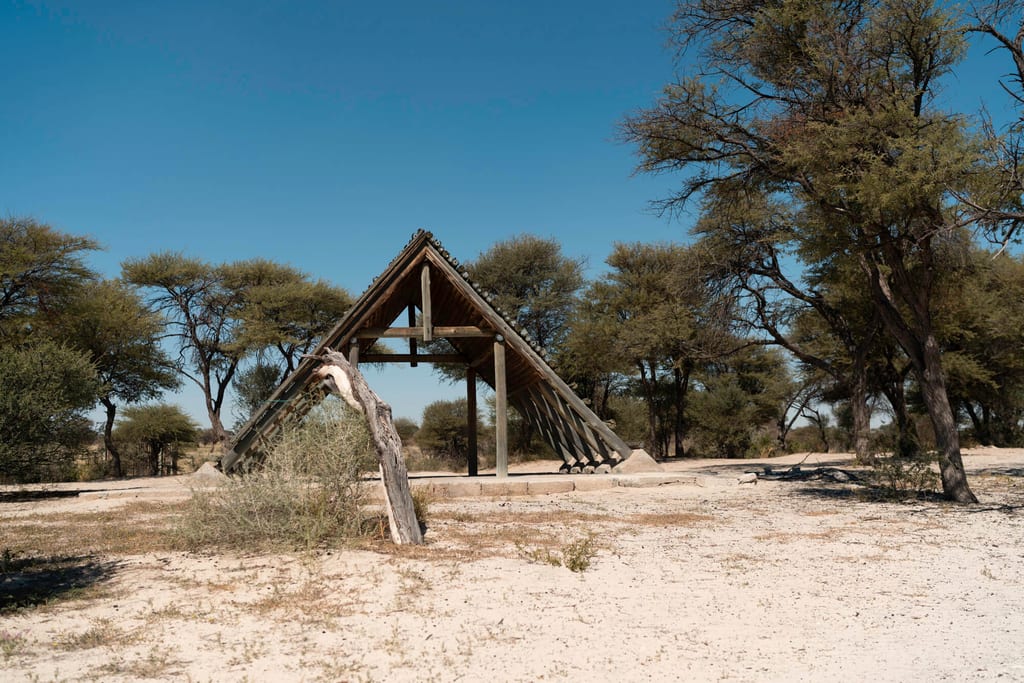 Dreieckige Holzüberdachung in sandiger Savannenlandschaft unter klarem blauem Himmel.