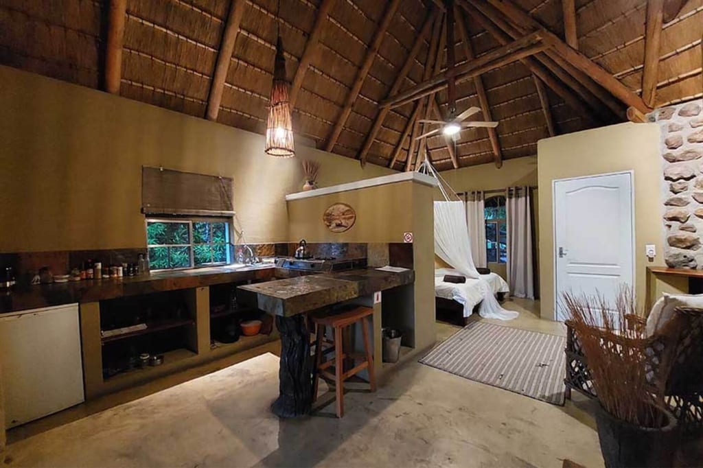Rustikale Lodge mit Strohdach, offener Küchenzeile und Bett mit Moskitonetz in warmem Licht.